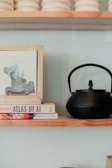 A shelf with a black teapot sits next to a small stack of books and a framed abstract painting. The books include titles on artificial intelligence, topped with a loose paper. There are stacked off-white ceramic bowls above.