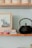A shelf with a black teapot sits next to a small stack of books and a framed abstract painting. The books include titles on artificial intelligence, topped with a loose paper. There are stacked off-white ceramic bowls above.