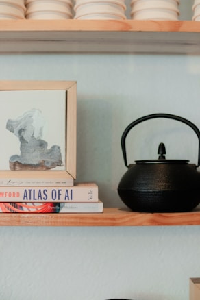 A shelf with a black teapot sits next to a small stack of books and a framed abstract painting. The books include titles on artificial intelligence, topped with a loose paper. There are stacked off-white ceramic bowls above.