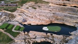 An aerial view of a unique golf course feature embedded in a rocky landscape. The central focus is a small, isolated green island surrounded by water, connected by a narrow bridge. Adjacent to the island, there are neatly maintained grassy tee areas and a rustic cabin or clubhouse amid the rocky terrain.