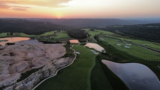 Aerial view of a golf course during sunset with green fairways, sand traps, and reflective water hazards. The landscape features a mix of rolling hills, rocky terrain, and dense forested areas in the background. The skyline is tinged with warm hues from the setting sun.