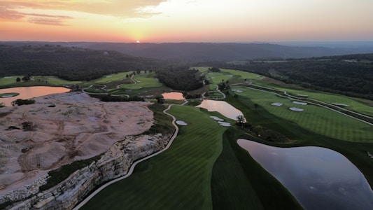 Aerial view of a golf course during sunset with green fairways, sand traps, and reflective water hazards. The landscape features a mix of rolling hills, rocky terrain, and dense forested areas in the background. The skyline is tinged with warm hues from the setting sun.