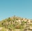 A picturesque hillside village comprised of rustic houses with terracotta roofs surrounded by lush greenery and tall cypress trees. The buildings are nestled among vibrant foliage under a clear blue sky, creating a serene Mediterranean atmosphere.