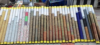 Rows of rolled up woven mats are on display, each with varying patterns and colors. The mats are neatly arranged side by side, with yellow tape wrapped around the ends. They all appear to be of similar size and are displayed on a tiled outdoor surface with additional rolls and items seen in the background.