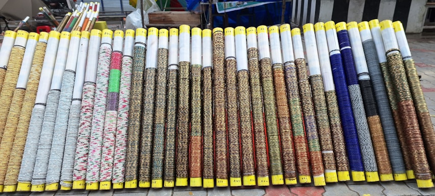 Rows of rolled up woven mats are on display, each with varying patterns and colors. The mats are neatly arranged side by side, with yellow tape wrapped around the ends. They all appear to be of similar size and are displayed on a tiled outdoor surface with additional rolls and items seen in the background.