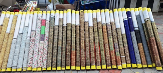 Rows of rolled up woven mats are on display, each with varying patterns and colors. The mats are neatly arranged side by side, with yellow tape wrapped around the ends. They all appear to be of similar size and are displayed on a tiled outdoor surface with additional rolls and items seen in the background.