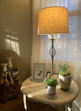A warmly lit room corner with a table lamp casting soft light. The table holds a framed botanical art piece and two potted succulent plants. Beside it, a small wooden stool features decorative candle holders and a bowl with tassels. Sunlight filters through sheer curtains, creating a cozy and inviting atmosphere.