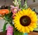A vibrant bouquet of flowers featuring a large sunflower with bright yellow petals, accompanied by pink snapdragons and an orange gerbera daisy. The bouquet includes greenery and a pink tag with text, celebrating a wedding day. The background shows a cozy indoor setting with framed photos and a white brick wall.