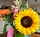 A vibrant bouquet of flowers featuring a large sunflower with bright yellow petals, accompanied by pink snapdragons and an orange gerbera daisy. The bouquet includes greenery and a pink tag with text, celebrating a wedding day. The background shows a cozy indoor setting with framed photos and a white brick wall.
