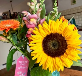 A vibrant bouquet of flowers featuring a large sunflower with bright yellow petals, accompanied by pink snapdragons and an orange gerbera daisy. The bouquet includes greenery and a pink tag with text, celebrating a wedding day. The background shows a cozy indoor setting with framed photos and a white brick wall.