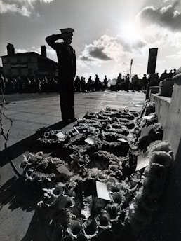 A silhouetted figure in uniform stands saluting beside a display of floral wreaths. The scene appears to be outdoors and possibly at a memorial or remembrance event. The gathering crowd in the background, set against a bright sky with clouds, suggests a formal or solemn occasion.