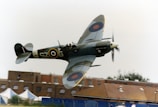 A vintage military aircraft in flight, flying close to a building. The aircraft has distinctive roundels on its wings, a propeller in motion, and markings on its body. The background shows a building with a brown roof, windows, and several tents.
