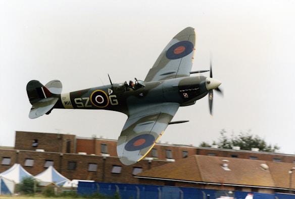 A vintage military aircraft in flight, flying close to a building. The aircraft has distinctive roundels on its wings, a propeller in motion, and markings on its body. The background shows a building with a brown roof, windows, and several tents.