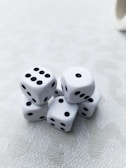 A cluster of six white dice with black dots is arranged on a light-textured surface. Each die displays a different number of dots, presenting a random assortment. The lighting is soft, creating subtle shadows and highlights on the dice.