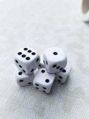 A cluster of six white dice with black dots is arranged on a light-textured surface. Each die displays a different number of dots, presenting a random assortment. The lighting is soft, creating subtle shadows and highlights on the dice.