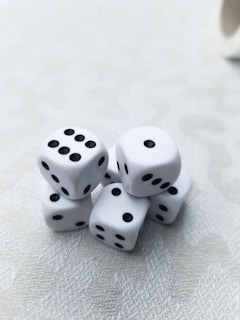A cluster of six white dice with black dots is arranged on a light-textured surface. Each die displays a different number of dots, presenting a random assortment. The lighting is soft, creating subtle shadows and highlights on the dice.
