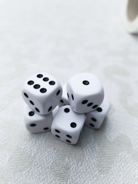 A cluster of six white dice with black dots is arranged on a light-textured surface. Each die displays a different number of dots, presenting a random assortment. The lighting is soft, creating subtle shadows and highlights on the dice.