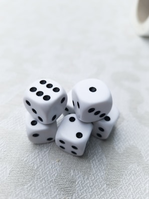 A cluster of six white dice with black dots is arranged on a light-textured surface. Each die displays a different number of dots, presenting a random assortment. The lighting is soft, creating subtle shadows and highlights on the dice.
