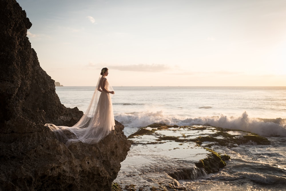 a woman in a wedding dress standing on a rock near the ocean