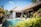 A tropical villa with a thatched roof is surrounded by lush greenery and vibrant plants. The villa features an inviting swimming pool with clear blue water in the foreground. Sunlight enhances the vivid colors of the foliage, and the architecture blends seamlessly with the natural surroundings. Two lounge chairs are located near the pool, providing a relaxing atmosphere.