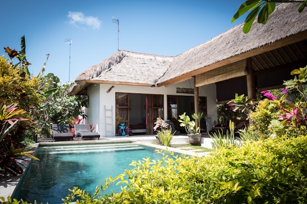 A tropical villa with a thatched roof is surrounded by lush greenery and vibrant plants. The villa features an inviting swimming pool with clear blue water in the foreground. Sunlight enhances the vivid colors of the foliage, and the architecture blends seamlessly with the natural surroundings. Two lounge chairs are located near the pool, providing a relaxing atmosphere.