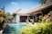 A tropical villa with a thatched roof is surrounded by lush greenery and vibrant plants. The villa features an inviting swimming pool with clear blue water in the foreground. Sunlight enhances the vivid colors of the foliage, and the architecture blends seamlessly with the natural surroundings. Two lounge chairs are located near the pool, providing a relaxing atmosphere.