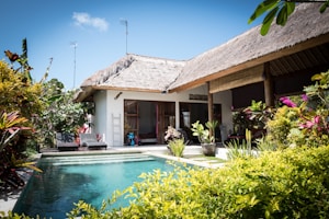 A tropical villa with a thatched roof is surrounded by lush greenery and vibrant plants. The villa features an inviting swimming pool with clear blue water in the foreground. Sunlight enhances the vivid colors of the foliage, and the architecture blends seamlessly with the natural surroundings. Two lounge chairs are located near the pool, providing a relaxing atmosphere.