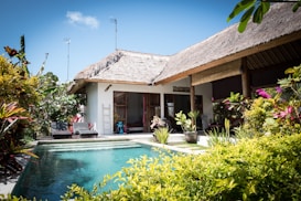 A tropical villa with a thatched roof is surrounded by lush greenery and vibrant plants. The villa features an inviting swimming pool with clear blue water in the foreground. Sunlight enhances the vivid colors of the foliage, and the architecture blends seamlessly with the natural surroundings. Two lounge chairs are located near the pool, providing a relaxing atmosphere.