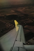 An airplane's wing is visible from a passenger's viewpoint, with a landscape of fields and a cluster of buildings below. The wingtip is marked with 'airBaltic' in green. The scene is captured during a sunset or sunrise, casting a warm, dim light over the landscape.