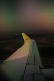 A view from an airplane window showing the aircraft's wing and a yellow tail fin with the logo 'airBaltic'. The landscape below is visible under soft, diffused lighting, possibly during sunset or sunrise, casting a warm glow over the scene.
