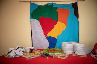 A colorful fabric map hangs on a wall above a table set up for a meal or event. On the table are stacks of white plates, utensils, and dishes, all arranged neatly. The table is covered with a red tablecloth and there are items like a bowl and decorative greenery.