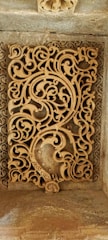 An intricate stone carving featuring ornate, swirling patterns of arabesque motifs. The design includes floral and geometric elements intricately intertwined, crafted in a rectangular panel set within a frame. The craftsmanship reflects detailed artistry in stonework.