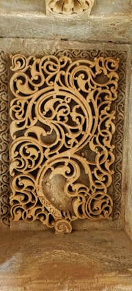 An intricate stone carving featuring ornate, swirling patterns of arabesque motifs. The design includes floral and geometric elements intricately intertwined, crafted in a rectangular panel set within a frame. The craftsmanship reflects detailed artistry in stonework.