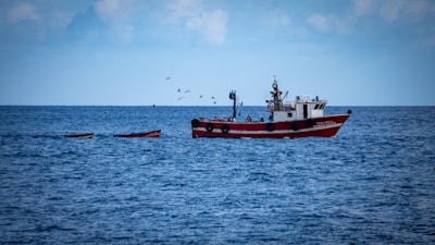A red and white fishing boat is sailing on a calm and vast blue ocean with several small boats nearby. A flock of birds hovers above the water, creating a serene maritime scene.