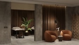 A modern and cozy interior space featuring two round, brown armchairs positioned beside a small round side table against a textured gray and wood-paneled wall. A decorative plant sits next to massage tables in a dimly lit area, adding an element of tranquility. Bamboo décor elements enhance the overall aesthetic.