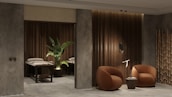 A modern and cozy interior space featuring two round, brown armchairs positioned beside a small round side table against a textured gray and wood-paneled wall. A decorative plant sits next to massage tables in a dimly lit area, adding an element of tranquility. Bamboo décor elements enhance the overall aesthetic.