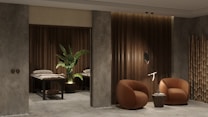 A modern and cozy interior space featuring two round, brown armchairs positioned beside a small round side table against a textured gray and wood-paneled wall. A decorative plant sits next to massage tables in a dimly lit area, adding an element of tranquility. Bamboo d&eacute;cor elements enhance the overall aesthetic.