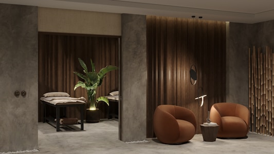 A modern and cozy interior space featuring two round, brown armchairs positioned beside a small round side table against a textured gray and wood-paneled wall. A decorative plant sits next to massage tables in a dimly lit area, adding an element of tranquility. Bamboo décor elements enhance the overall aesthetic.