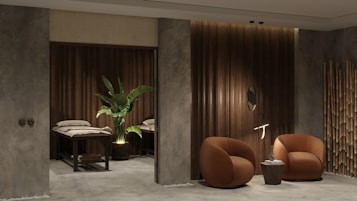 A modern and cozy interior space featuring two round, brown armchairs positioned beside a small round side table against a textured gray and wood-paneled wall. A decorative plant sits next to massage tables in a dimly lit area, adding an element of tranquility. Bamboo d&eacute;cor elements enhance the overall aesthetic.