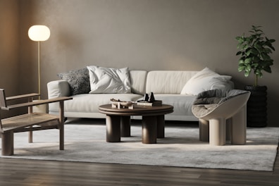 A modern living room with a beige sofa adorned with plush pillows and a patterned cushion. The room features a round wooden coffee table with decorative items. A stylish chair with a woven seat and a distinctive curved chair are positioned on a light gray rug. A soft-lit floor lamp stands on the left, and a lush green plant in a textured black pot is on the right, against a neutral wall.