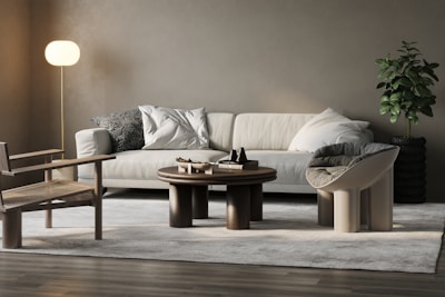 A modern living room with a beige sofa adorned with plush pillows and a patterned cushion. The room features a round wooden coffee table with decorative items. A stylish chair with a woven seat and a distinctive curved chair are positioned on a light gray rug. A soft-lit floor lamp stands on the left, and a lush green plant in a textured black pot is on the right, against a neutral wall.
