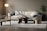 A modern living room with a beige sofa adorned with plush pillows and a patterned cushion. The room features a round wooden coffee table with decorative items. A stylish chair with a woven seat and a distinctive curved chair are positioned on a light gray rug. A soft-lit floor lamp stands on the left, and a lush green plant in a textured black pot is on the right, against a neutral wall.