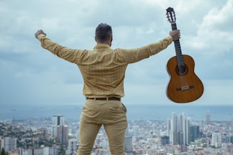 A person dressed in beige attire stands on a hillside overlooking a sprawling cityscape. Arms are outstretched, holding an acoustic guitar in one hand. The view includes high-rise buildings and a distant ocean horizon under a cloudy sky.