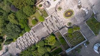 An aerial view of a formal garden with symmetrical designs, pathways, and green hedges. The space includes circular and rectangular flowerbeds, a wide staircase, and a paved plaza. Several people are visible walking through the area, surrounded by lush greenery.