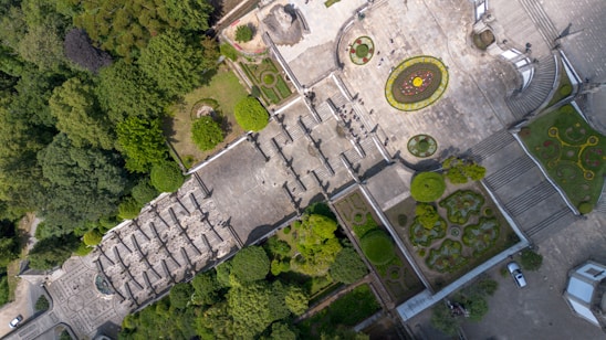 An aerial view of a formal garden with symmetrical designs, pathways, and green hedges. The space includes circular and rectangular flowerbeds, a wide staircase, and a paved plaza. Several people are visible walking through the area, surrounded by lush greenery.