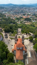 An aerial view of a baroque-style church with a red-tiled roof, surrounded by lush greenery and gardens. There is a wide staircase leading up to the church, with a circular flower bed in front. In the background, a cityscape extends into the distance, bordered by rolling hills under a partly cloudy sky.