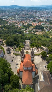 An aerial view of a baroque-style church with a red-tiled roof, surrounded by lush greenery and gardens. There is a wide staircase leading up to the church, with a circular flower bed in front. In the background, a cityscape extends into the distance, bordered by rolling hills under a partly cloudy sky.