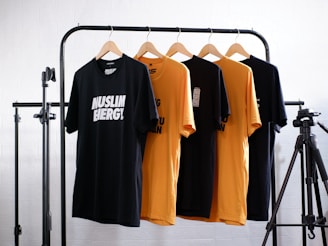 Four t-shirts, two black and two yellow, are hanging on wooden hangers from a black metal clothes rack. The shirts have different texts printed on them. There is a camera tripod on the right side of the frame. The background is a white brick wall.