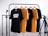 Four t-shirts, two black and two yellow, are hanging on wooden hangers from a black metal clothes rack. The shirts have different texts printed on them. There is a camera tripod on the right side of the frame. The background is a white brick wall.