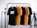 Four t-shirts, two black and two yellow, are hanging on wooden hangers from a black metal clothes rack. The shirts have different texts printed on them. There is a camera tripod on the right side of the frame. The background is a white brick wall.